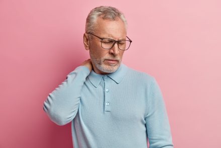 Image of dissatisfied elderly man touches neck, suffers from pain, closes eyes from displeasure, wears optical glasses, casual jumper, poses against pink background, feels unwell and overworked